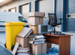 Commercial waste including cardboard boxes, paper, plastic bottles, broken office furniture and old electronics stacked outside a workplace with a recycling bin and clearance van in the background